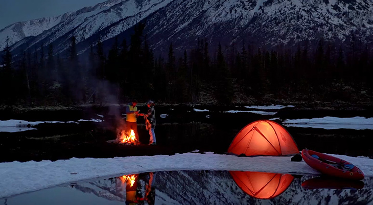 Zwei Personen stehen bei einem Lagerfeuer in einer verschneiten, bergigen Landschaft. Neben ihnen steht ein beleuchtetes orangefarbenes Zelt, das sich im Wasser spiegelt. Rechts im Bild liegt ein aufblasbares rotes Boot am Ufer. Im Hintergrund sind verschneite Berge und dunkle Bäume zu sehen, es ist bereits dämmrig oder Nacht.