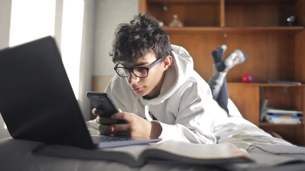 Eine Person in weißem Hoodie und Brille liegt auf dem Bauch, nutzt ein Smartphone. Daneben sind ein Laptop und ein offenes Buch. Im Hintergrund ein Holzregal.