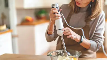 Person in brauner Schürze und grauem Pullover mixt mit weiß-silbernem Stabmixer weiße und gelbe Zutaten in einer Glasschüssel auf Holztisch in Küche.