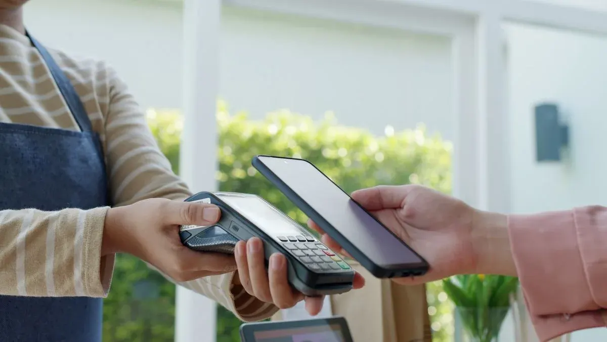 Eine Person hält ein Zahlterminal, während eine andere Person ein Smartphone für kontaktloses Bezahlen darüber hält. Im Hintergrund sind Fenster und Pflanzen zu sehen.
