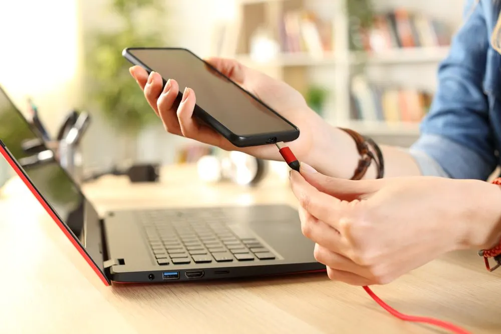 Person verbindet ein Smartphone mit einem Ladekabel an einen Laptop.