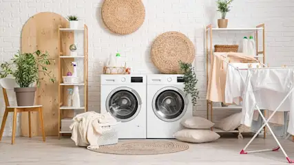 Heller Waschraum: Zwei weiße Waschmaschinen, Regale mit Pflanzen/Reinigern. Wäschekorb, Trockenständer mit Kleidung, Strohdekorationen, Holzboden.