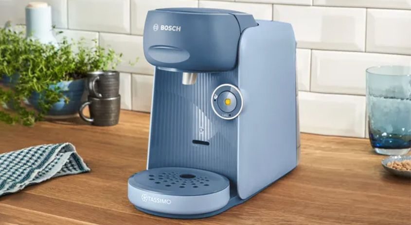 Auf einer Küchentheke steht eine blaue Bosch Tassimo Kaffeemaschine neben einem Glas und zwei gestapelten Tassen.