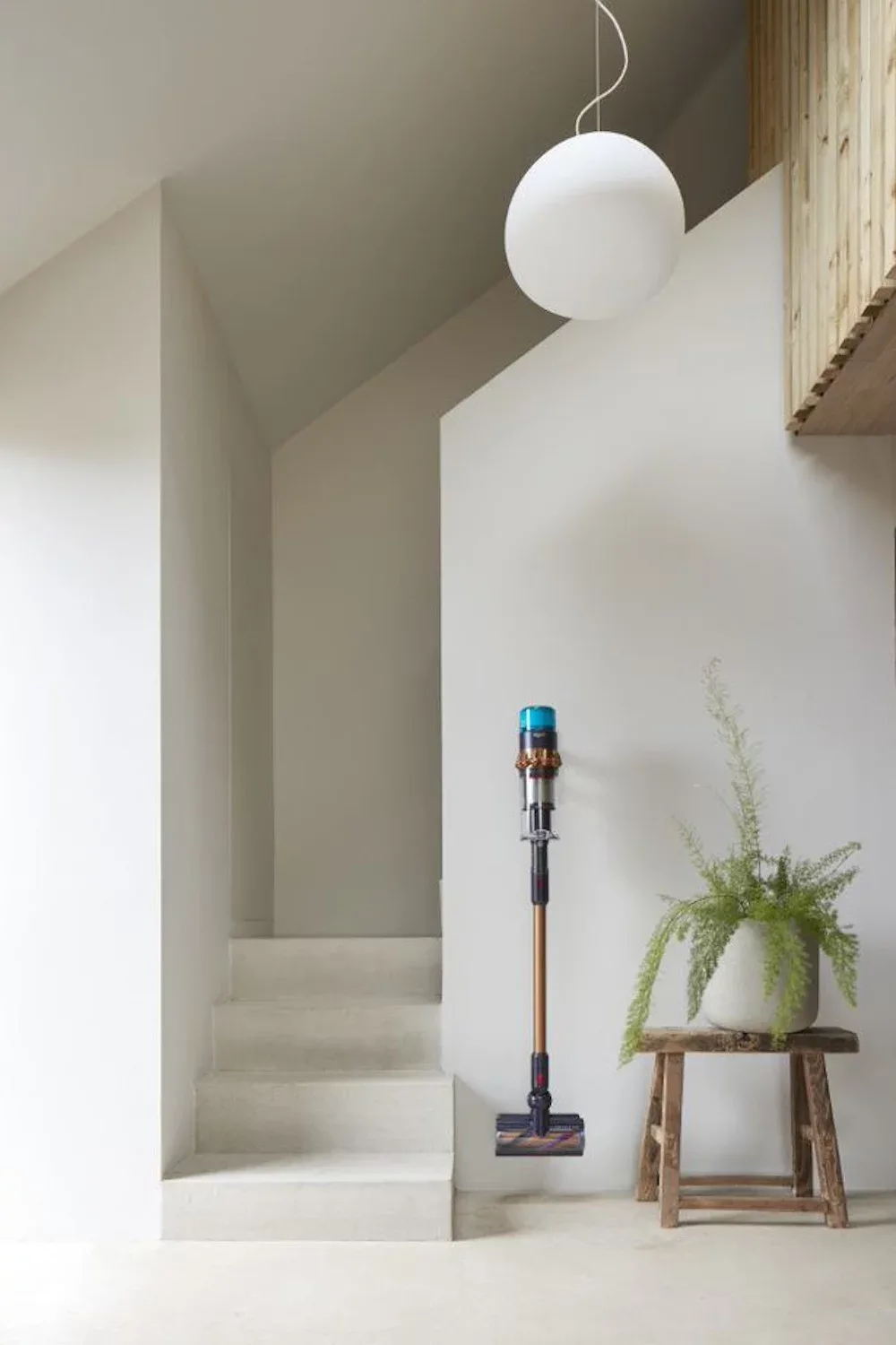 Minimalistischer Raum mit grauen Treppen, weißer Kugel-Pendelleuchte. Blauer Dyson-Staubsauger an Wand, daneben Holzhocker mit Pflanze.