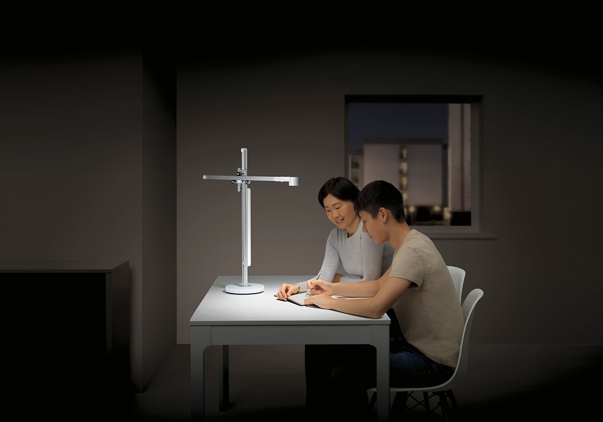 Zwei Personen sitzen an einem Tisch unter einer Lampe. Durch ein Fenster ist ein Gebäude sichtbar.