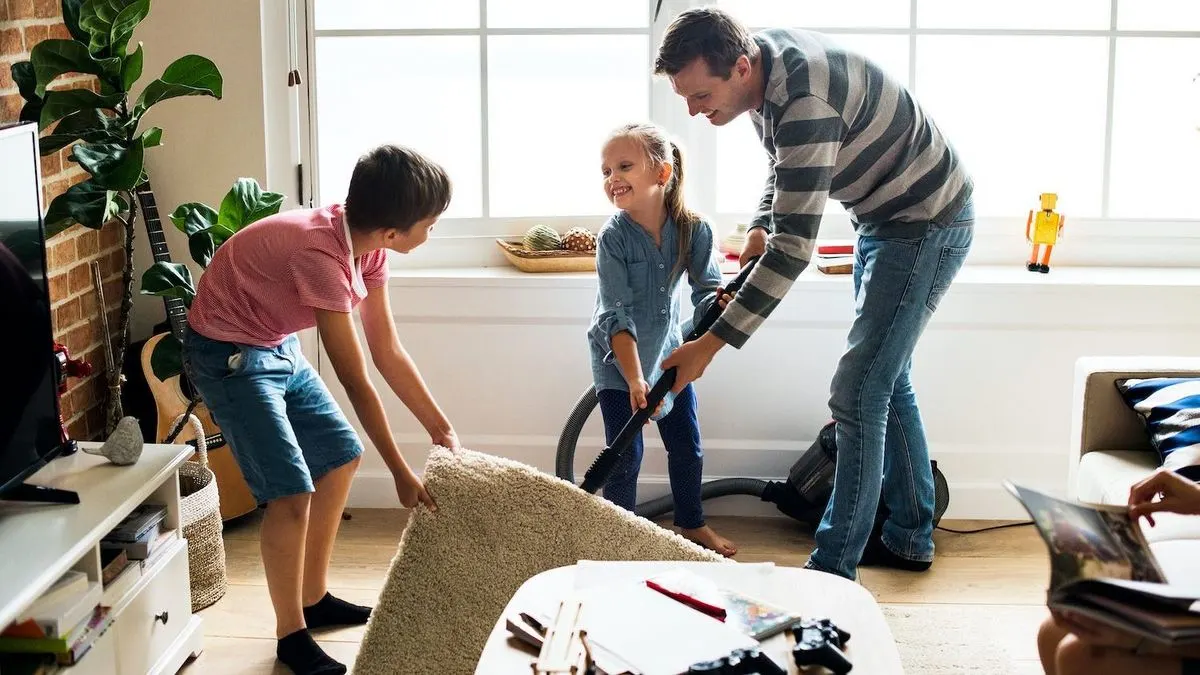 Vater und zwei Kinder putzen. Der Junge hebt einen beigen Teppich, das Mädchen saugt. Im Hintergrund sind Fenster, eine Pflanze und eine Gitarre.