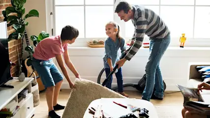 Vater und zwei Kinder putzen. Der Junge hebt einen beigen Teppich, das Mädchen saugt. Im Hintergrund sind Fenster, eine Pflanze und eine Gitarre.