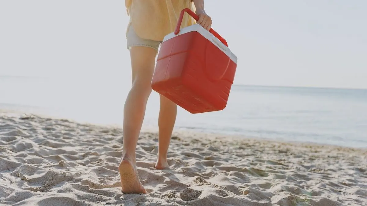 Person trägt rote Kühlbox barfuß am Strand entlang.