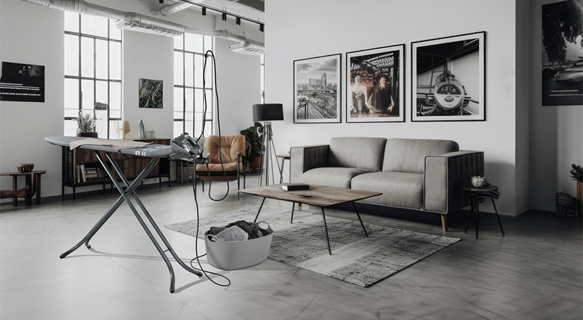 Ein Wohnzimmer mit Bügelbrett, grauem Sofa, Holztisch und gerahmten Bildern an der Wand. Wäschekorb im Vordergrund.