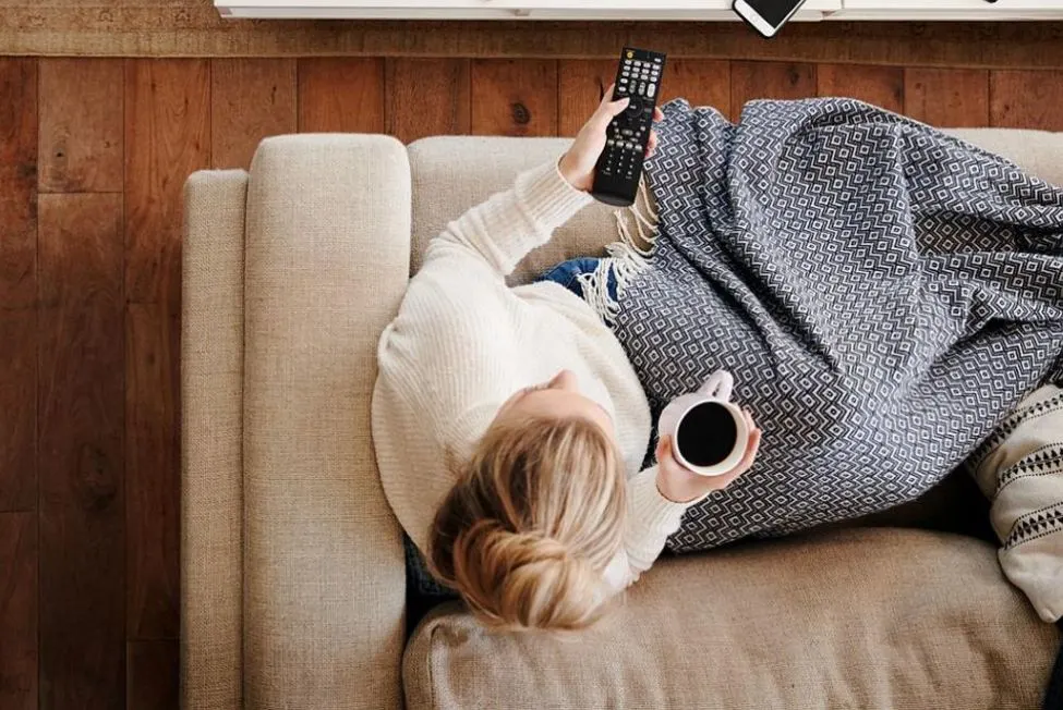 Ansicht einer Person auf dem Sofa von oben mit Fernbedienung und Kaffee in Haenden