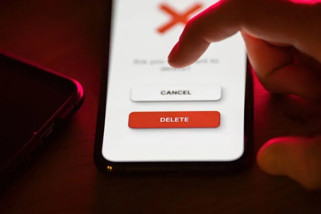 Finger tippt auf Smartphone-Bildschirm mit rotem X, weißem "CANCEL"- und rotem "DELETE"-Button. Daneben ein dunkles Smartphone.