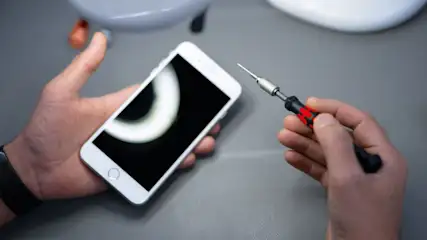 Hände halten ein weißes Smartphone und einen schwarzen Präzisionsschraubendreher mit rotem Muster. Grauer Tischhintergrund.