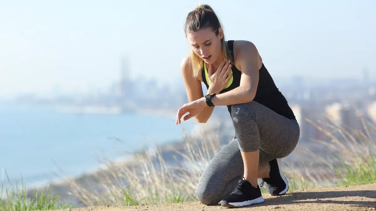 Eine erschöpfte Frau in Sportkleidung kniet beim Joggen auf einem Weg mit Blick auf eine Stadt und das Meer im Hintergrund.