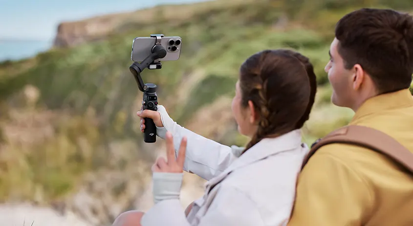 Hand hält Gimbal mit Smartphone vor Küstenlandschaft. Düne, Meer und Klippen sind im Hintergrund sichtbar.