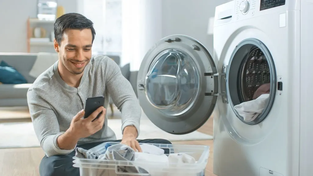 Mann sitzt vor einer offenen Waschmaschine und nutzt ein Smartphone