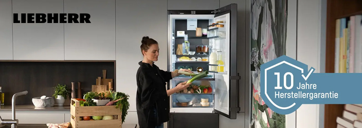 Moderne Küche mit geöffnetem Liebherr Kühl-Gefrierschrank in Edelstahl-Optik. Das Gerät zeigt gut organisierte Fächer mit verschiedenen Lebensmitteln. Prominente Werbeelemente zeigen "10 Jahre Herstellergarantie" und "Gültig auf alle Haushaltsgeräte 01.06. - 31.10.2026 (Kaufdatum)". Die Küche hat helle Holzfronten und eine warme, einladende Atmosphäre.