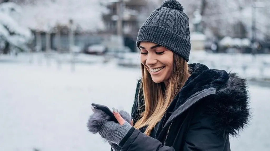 Frau mit grauer Muetze und Handschuhen benutzt ein Smartphone draussen im Schnee
