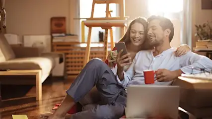 Ein lächelndes Paar sitzt auf Holzboden in einem sonnigen Raum. Die Frau hält Handy und grüne Tasse, der Mann eine rote Tasse; ein Laptop liegt daneben.