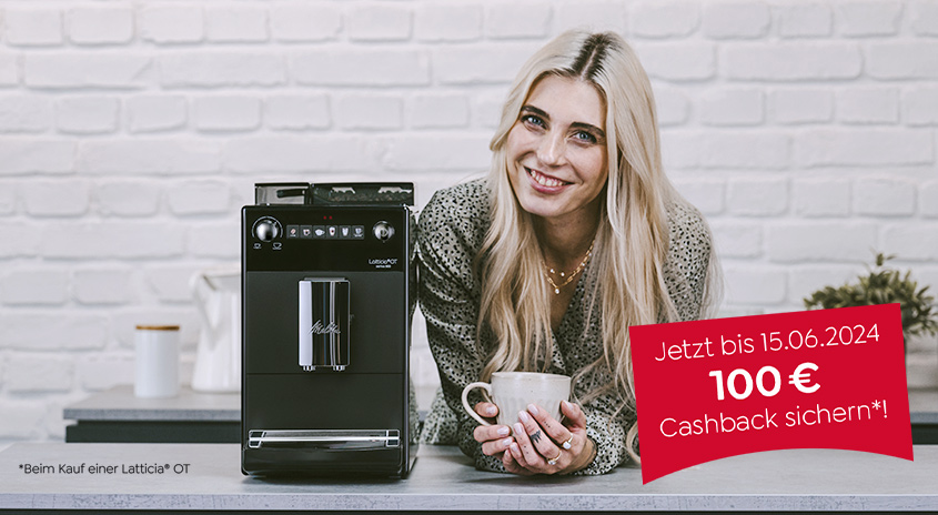 Eine schwarze Melitta-Kaffeemaschine steht auf einer grauen Arbeitsplatte vor einer weißen Ziegelwand. Ein rotes Schild wirbt mit 100 € Cashback.