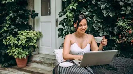 Lachende Frau tippt am Laptop an dunklem Tisch, hält weiße Tasse. Im Hintergrund grüne Pflanzen, weiße Tür, orange Blumen.