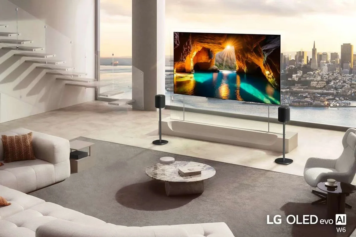 Weitläufiges, helles Wohnzimmer mit freistehendem, großem LG OLED-Fernseher auf schlankem Ständer, der eine leuchtende Meeresgrotte zeigt; zwei schlanke Lautsprecher auf Ständern; helle Polstermöbel, runder Marmorcouchtisch und ein moderner Sessel; große Glasfenster mit Blick auf eine Stadtsilhouette und eine Treppe mit Glasgeländer im Hintergrund. Logo „LG OLED evo AI W6“ sichtbar.