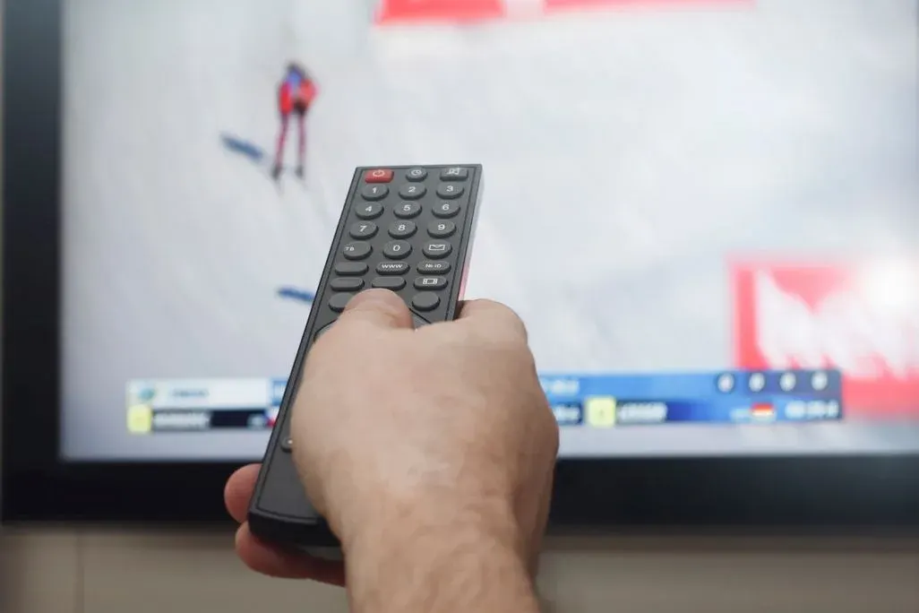 Hand hält eine TV‑Fernbedienung vor einem Fernseher, auf dem eine Skisport‑Übertragung mit Athlet und Ergebnisleiste zu sehen ist.