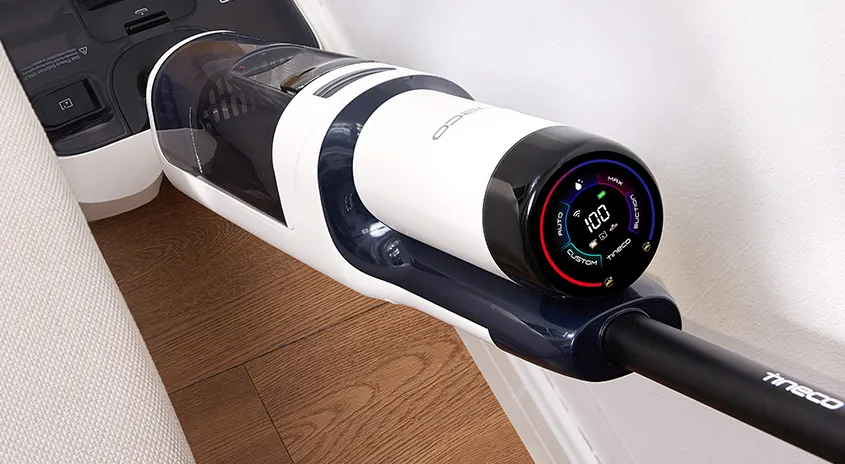 Ein kabelloser Staubsauger steht auf einem Holzboden neben einem cremefarbenen Sofa. Der Staubsauger ist weiß und schwarz mit einem digitalen Display.