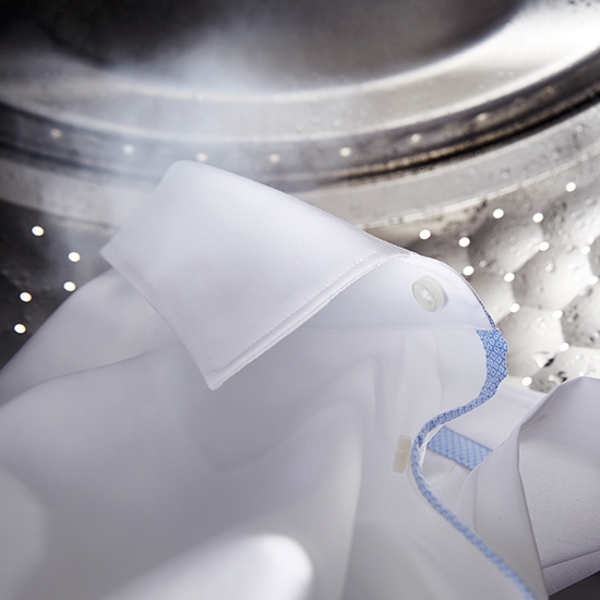 Weißes Hemd mit blauer Naht in einer silbernen Waschmaschine mit Wasserdampf.