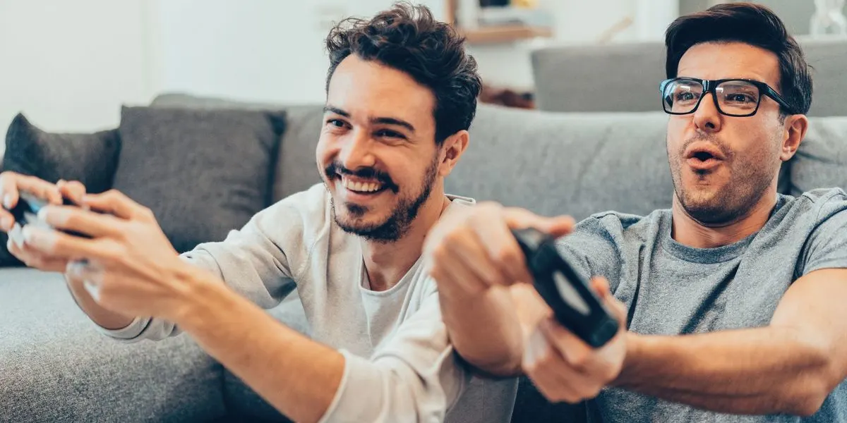 Zwei Männer sitzen auf einem grauen Sofa und spielen Videospiele mit schwarzen Controllern. Einer lacht, der andere mit Brille hat den Mund offen.