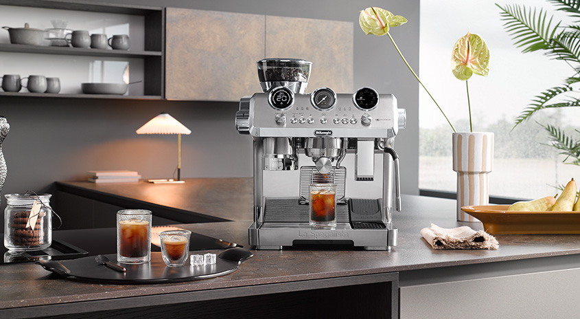 Eine silberne Espressomaschine auf einer Küchentheke, umgeben von Gläsern mit Kaffee, Keksen und einer Vase mit Blumen.