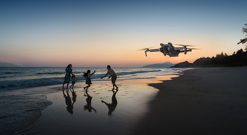 Eine Drohne schwebt am Strand über dem Meer, während die Sonne untergeht. Der Himmel ist blau und orange.