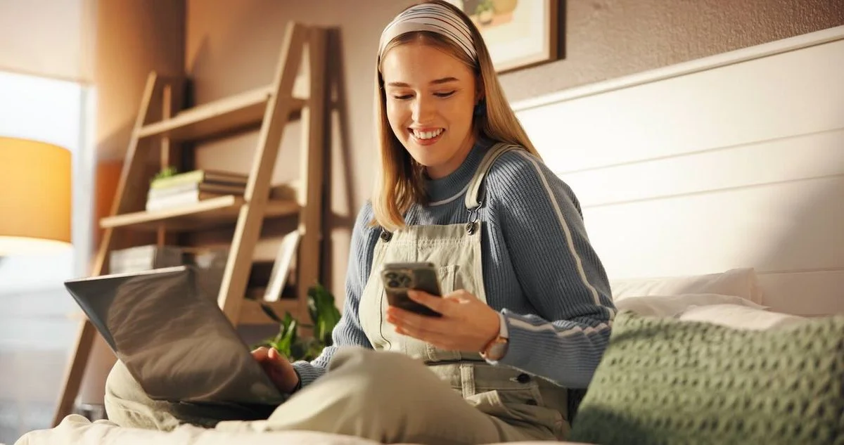 Junge Frau mit Laptop und Smartphone auf dem Bett, gemütliches Homeoffice-Szenario.