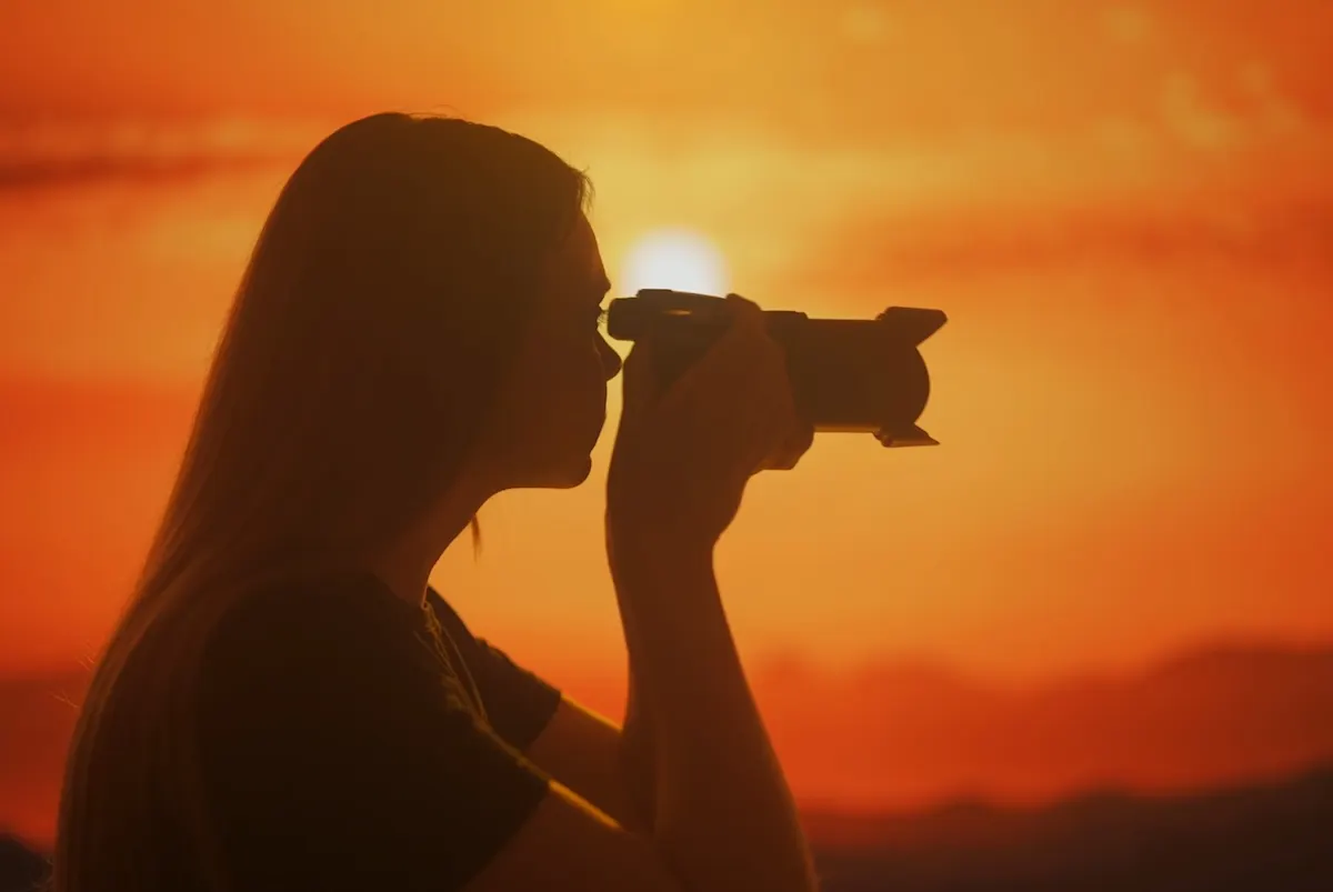 Die Silhouette einer Frau, die sich eine Kamera ans Auge hält vor einem orange-roten Sonnenuntergang.