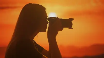 Die Silhouette einer Frau, die sich eine Kamera ans Auge hält vor einem orange-roten Sonnenuntergang.