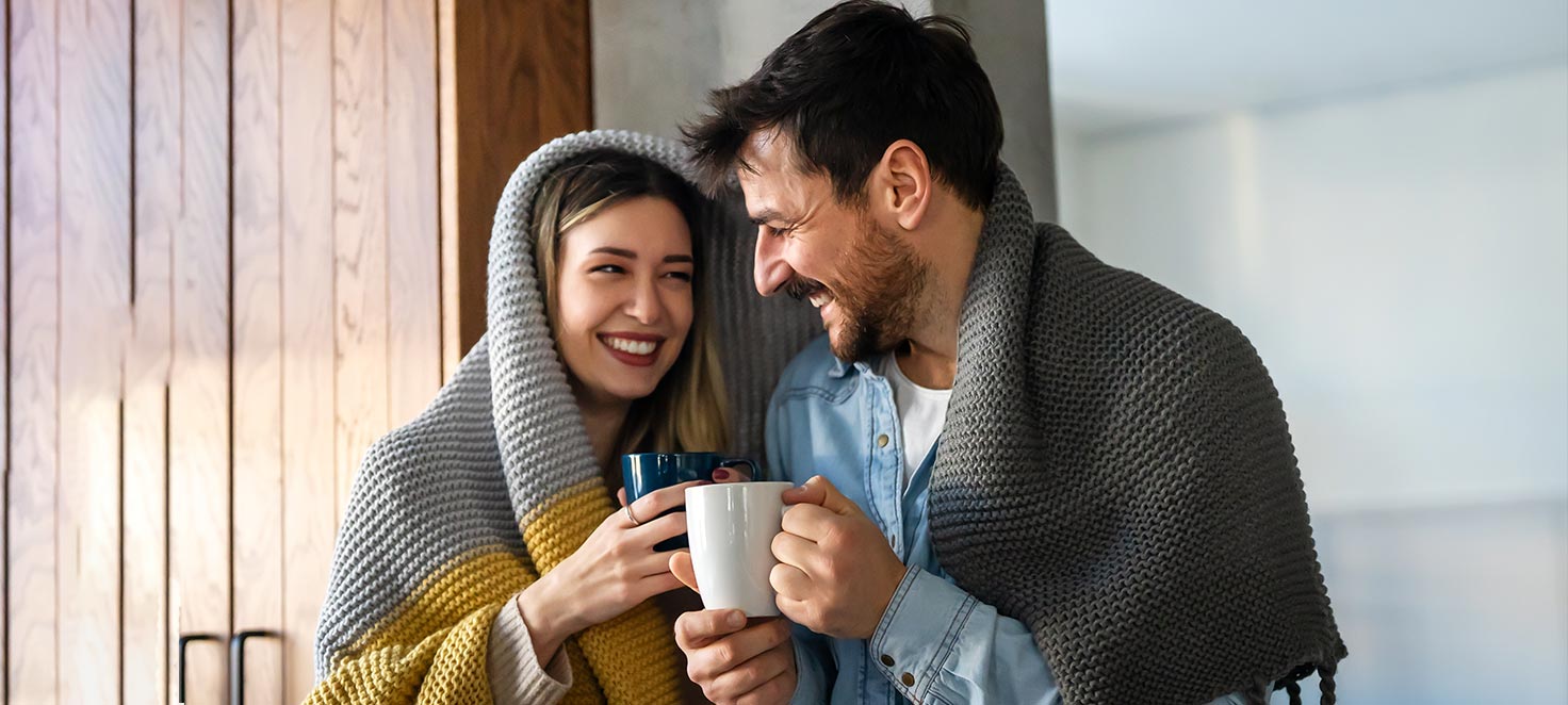 Ein Paar, eingewickelt in Decken, hält Tassen vor einer Holzwand. Die Decke ist grau und gelb.