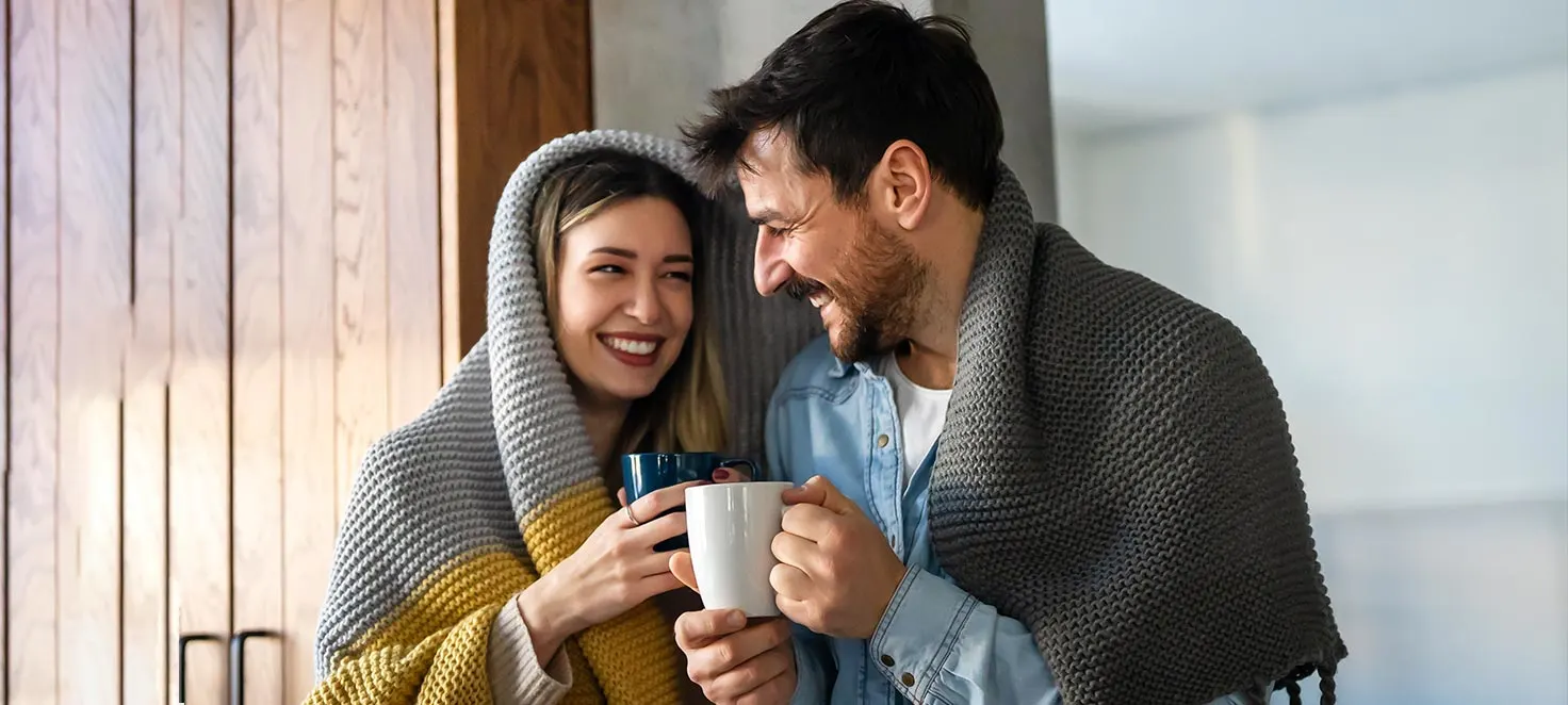 Ein Paar, eingewickelt in Decken, hält Tassen vor einer Holzwand. Die Decke ist grau und gelb.