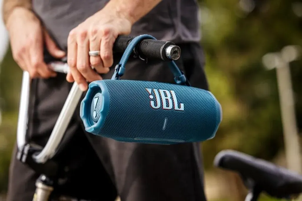 Eine Person hält ein Fahrrad am Lenker, an dem ein blauer JBL‑Bluetooth-Lautsprecher mit Trageschlaufe befestigt ist; im Hintergrund unscharfe, grüne Outdoor-Umgebung.