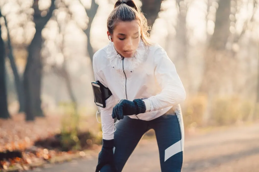 Frau macht Pause beim Joggen im Park und traegt Handschuhe sowie Smartwatch