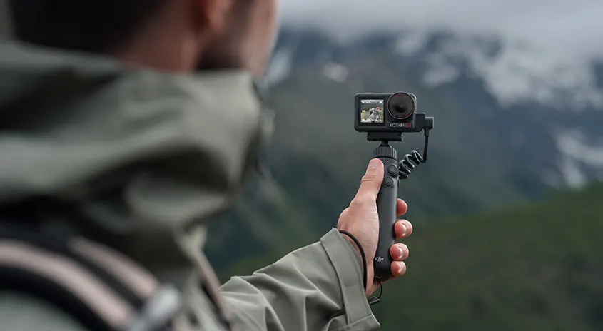 Eine Person hält eine schwarze Action-Kamera auf einem Stativ vor einer Berglandschaft.
