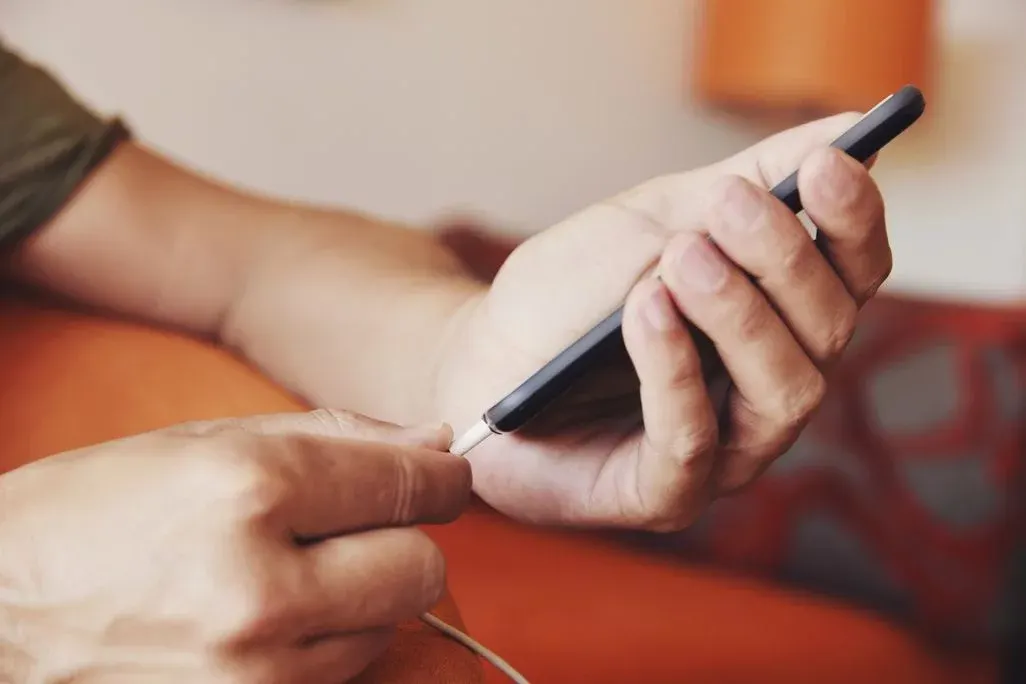Zwei Hände halten ein Smartphone, während ein Ladekabel angeschlossen wird – im Hintergrund ein orangefarbenes Sofa.
