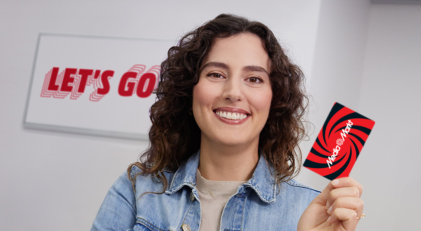 Eine Person hält eine rote MediaMarkt-Karte vor einem weißen Hintergrund mit dem Text „LET'S GO“.