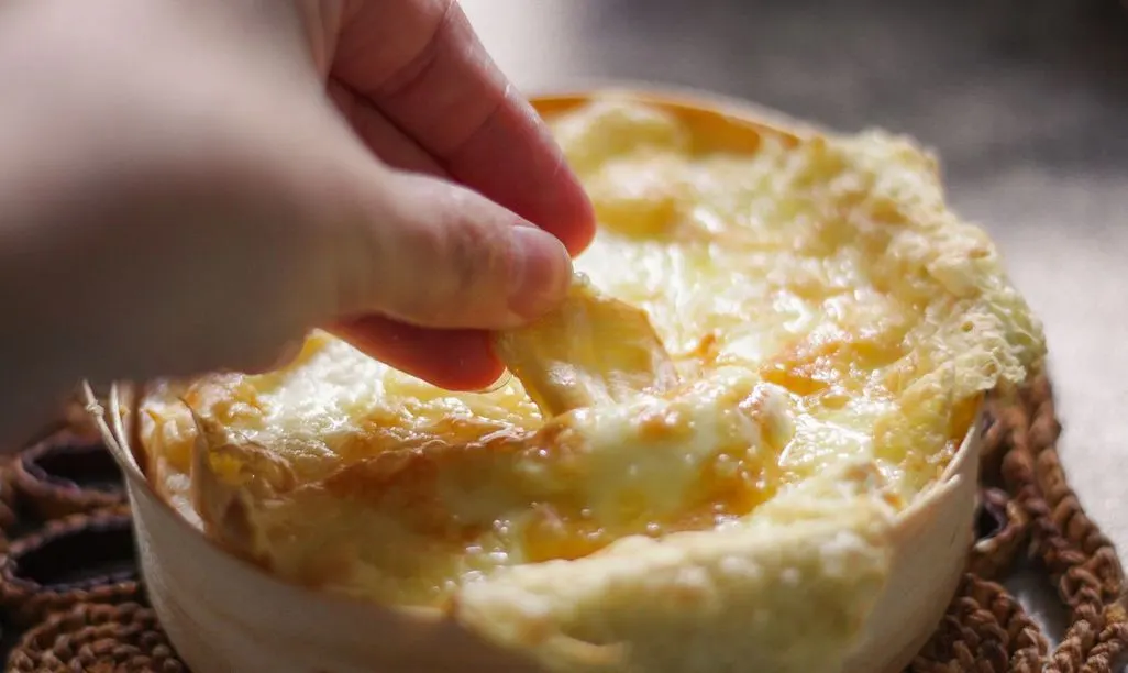 Eine Hand zieht ein Stück aus geschmolzenem, goldbraunen Käse, der Fäden zieht, aus einer hellen Schale.