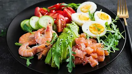 Bunt angerichteter Low-Carb-Teller mit Garnelen, Lachs, Avocado, Ei, Gurke, Tomaten und Rucola auf schwarzem Teller.