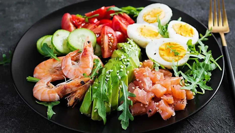 Bunt angerichteter Low-Carb-Teller mit Garnelen, Lachs, Avocado, Ei, Gurke, Tomaten und Rucola auf schwarzem Teller.
