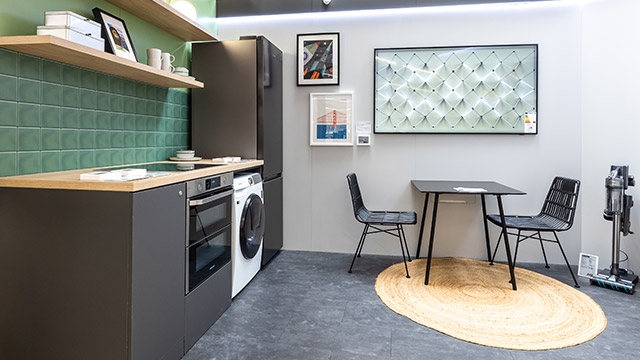 Eine Küche mit Geräten, darunter Kühlschrank, Waschmaschine und Ofen. Es gibt einen Tisch mit zwei Stühlen und einen runden Teppich.