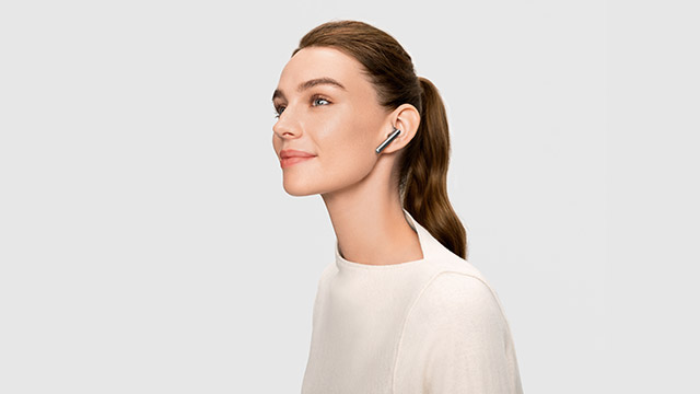 Eine Frau mit Ohrhörern vor einem hellen Hintergrund. Sie trägt ein helles Oberteil und ihr Haar ist zu einem Pferdeschwanz gebunden.