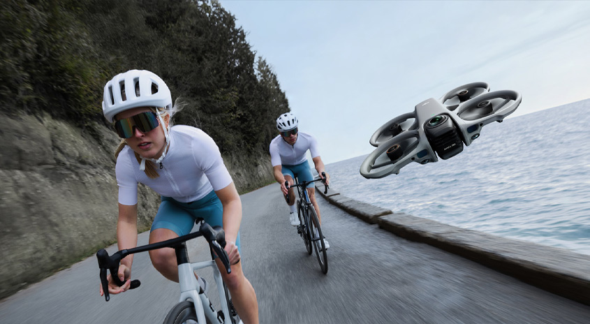 Drohne filmt zwei Radsportler auf schmaler Küstenstraße am Wasser, dynamische Fahrtaufnahme in Kurve mit Fokus auf Action und Umgebung.