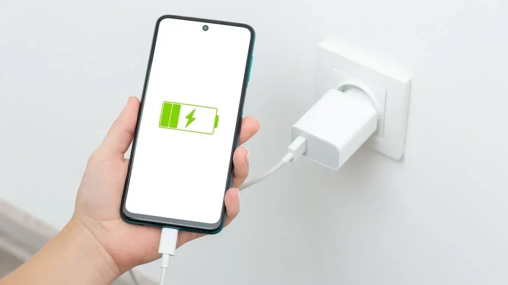 Hand hält Smartphone mit grünem Ladesymbol. Weißes Kabel verbindet Telefon mit weißem Netzteil in Steckdose.