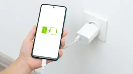 Hand hält Smartphone mit grünem Ladesymbol. Weißes Kabel verbindet Telefon mit weißem Netzteil in Steckdose.