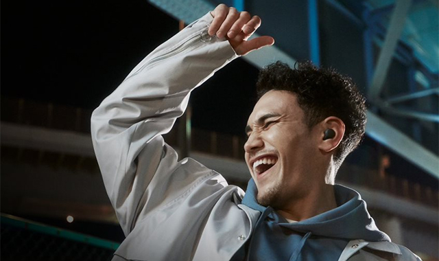 Ein lachender Mann mit Kopfhörern, blauem Hoodie und hellem Jackett hebt eine Hand. Im Hintergrund sind Gebäude zu sehen.